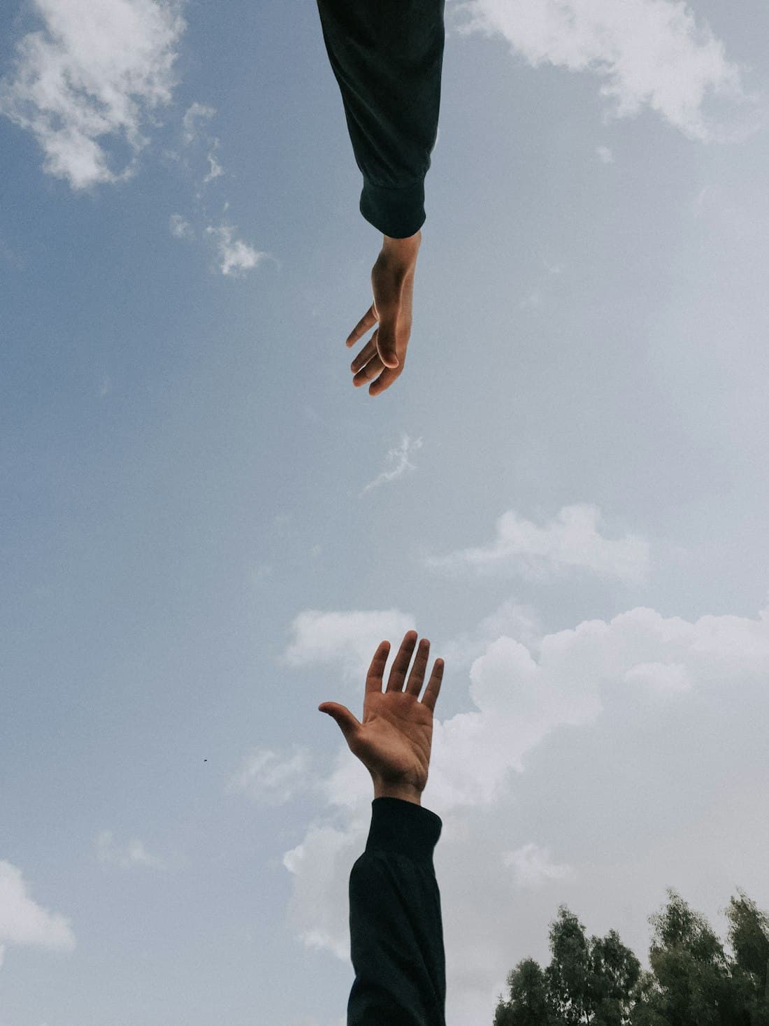Two people reaching a hand towards one another against an open sky.