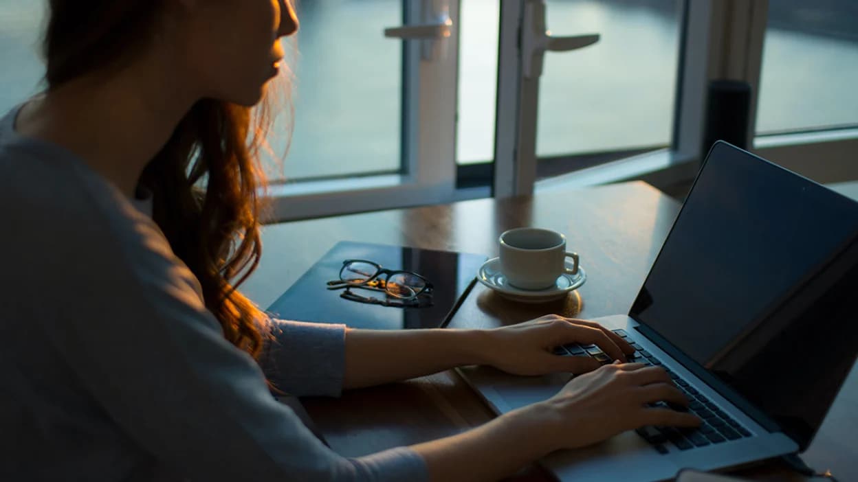 A woman working on a laptop at a table by a window, her glasses and a cup of coffee beside her.